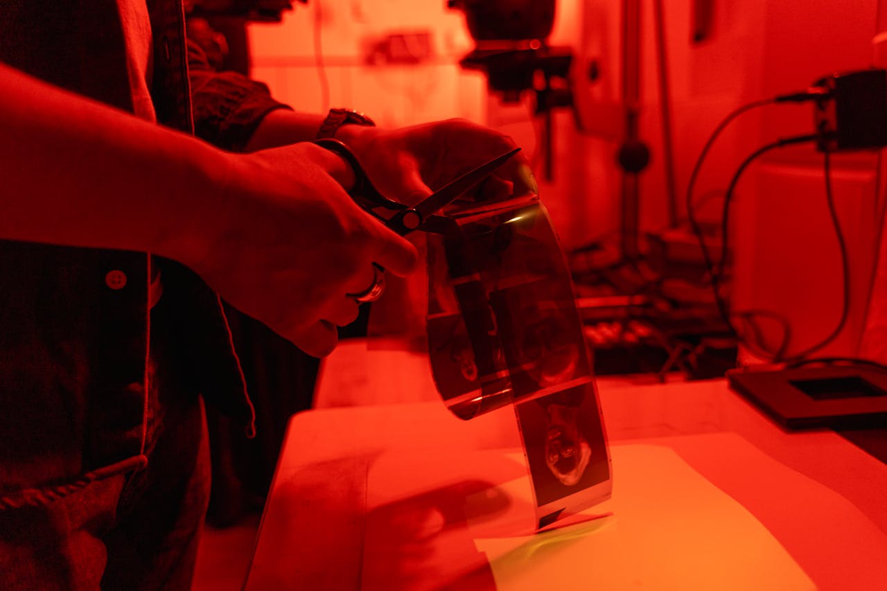 A person carefully cuts film negatives in a red-lit darkroom using scissors.